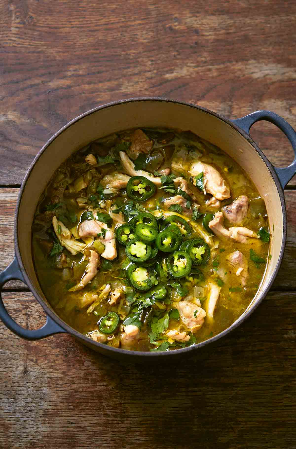 A large blue Dutch oven filled with green chicken chili, topped with sliced jalapenos.