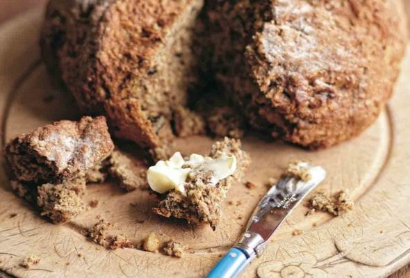 A loaf of Irish soda bread on a brown plate with a chunk cut out and smeared with butter.