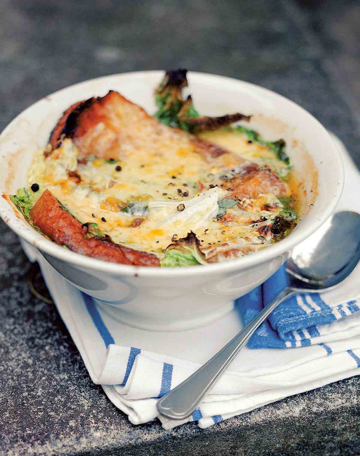 A white bowl filled with Italian kale soup, topped with ciabatta and melted cheese, with a spoon resting on the side.