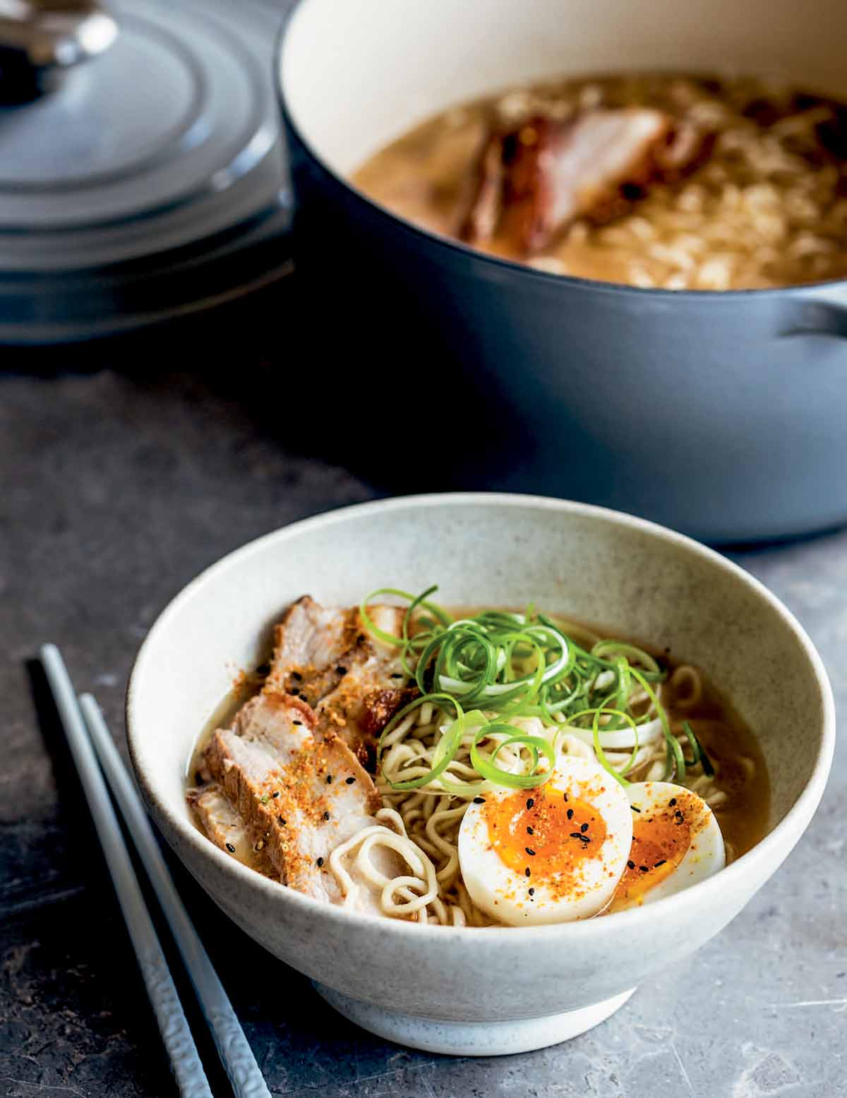 A white bowl filled with pork belly ramen, two egg halves, and scallions with a Dutch oven in the background and a pair of chopsticks lying beside the bowl.