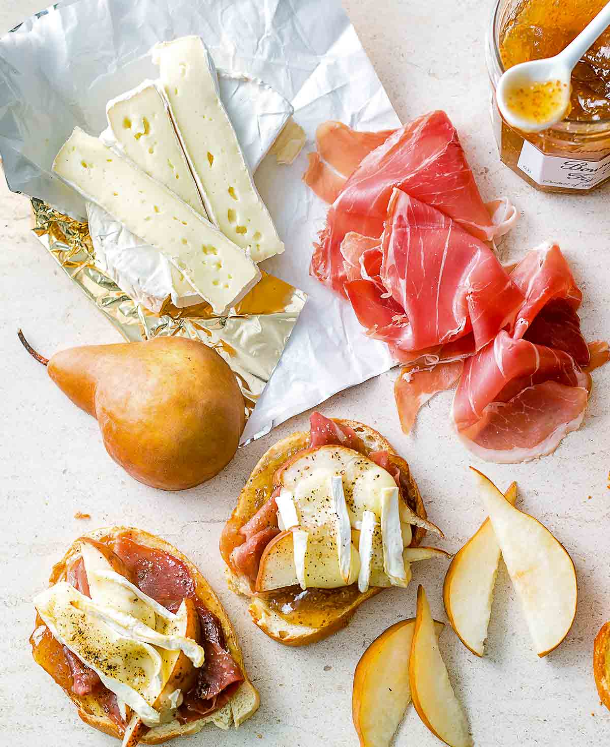 Two pieces of prosciutto, pear, and Brie toasts with slices of Brie, pear, prosciutto, and a jar of fig jam beside the toasts.
