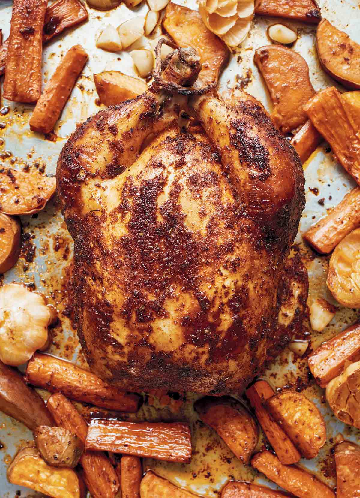 A whole roast chicken rubbed with paprika on a baking sheet with carrots and sweet potatoes.