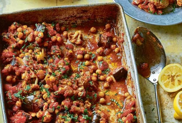 A metal baking tray filled with sheet pan chickpea curry with a spoon, two lemon halves, and a bowl of Greek yogurt beside it.