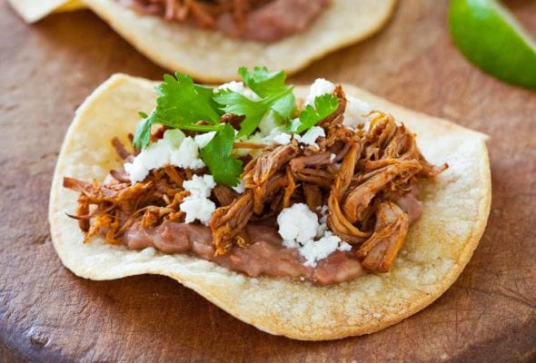 Two corn tortillas topped with slow cooker shredded pork, cheese, and cilantro on a wooden board with some lime wedges beside.