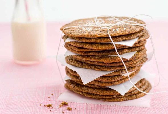 A stack of thin and crisp chocolate chip cookies with pieces of parchment in between, tied up with string and a glass bottle of milk in the background.