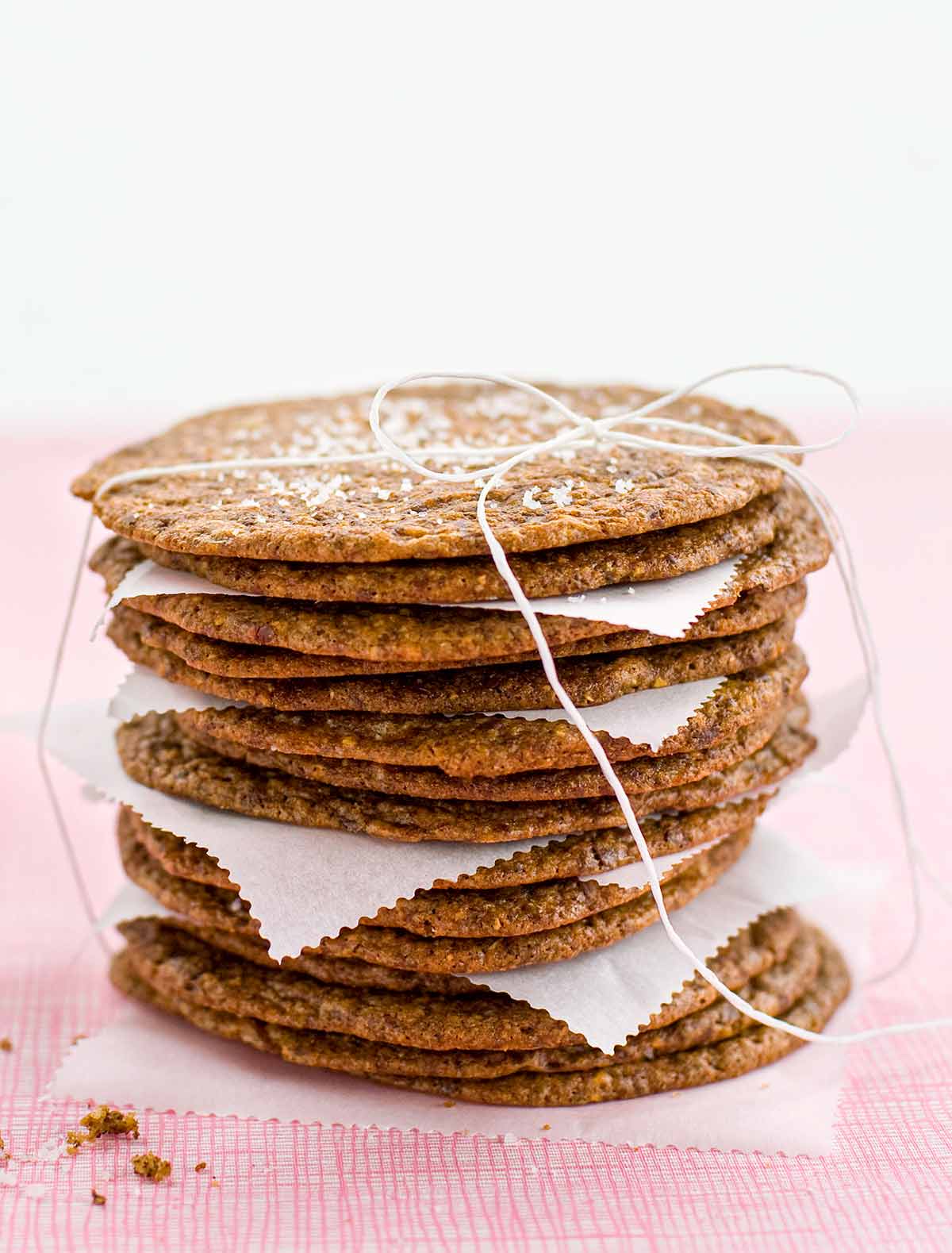 A stack of thin and crisp chocolate chip cookies with pieces of parchment in between, tied up with string and a glass bottle of milk in the background.