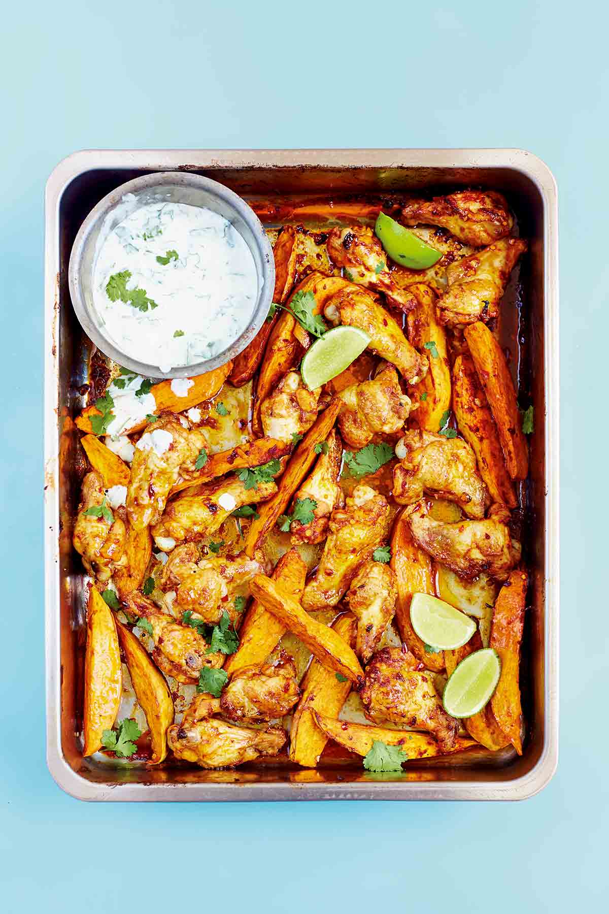 A metal tray filled with chipotle chicken wings, sweet potato wedges, and a Greek yogurt dipping sauce with lime wedges and cilantro leaves.