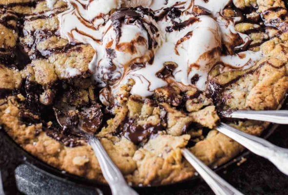 A chocolate chunk skillet cookie topped with ice cream and chocolate sauce in a cast-iron skillet with four spoons resting in the cookie.