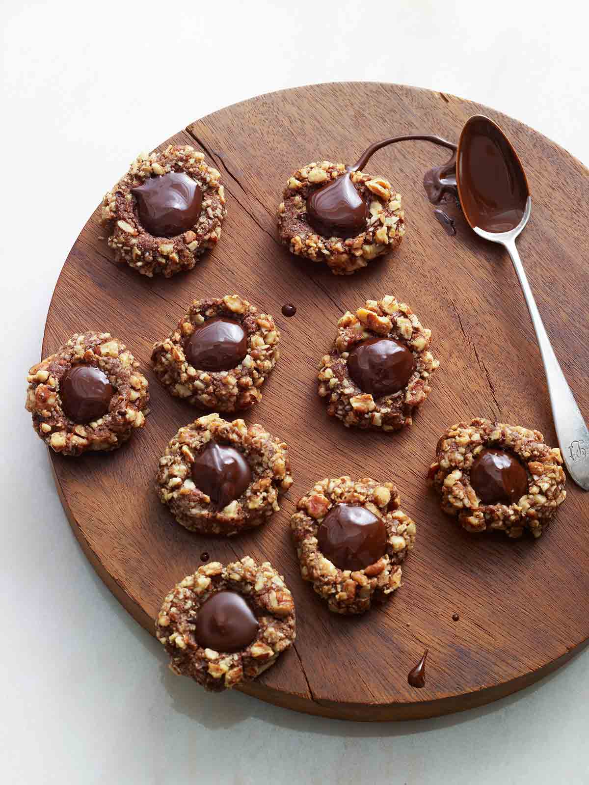 Nine chocolate thumbprint cookies and a chocolate covered spoon on a round wooden board.
