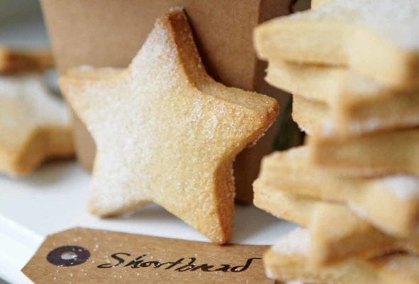 A stack of star-shaped cutout classic shortbread cookies, lightly dusted with sugar, and a brown takeout container and a gift tag