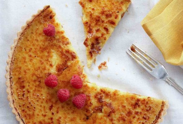 A partially sliced coconut crème brûlée tart topped with raspberries on a white linen cloth with a fork and napkin beside it.