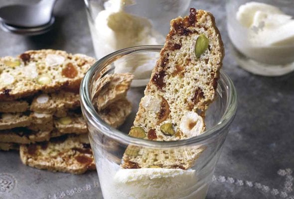 Three glasses with fig, apricot, and pistachio biscotti resting inside or on top of two of them and a stack of biscotti beside the glassware.