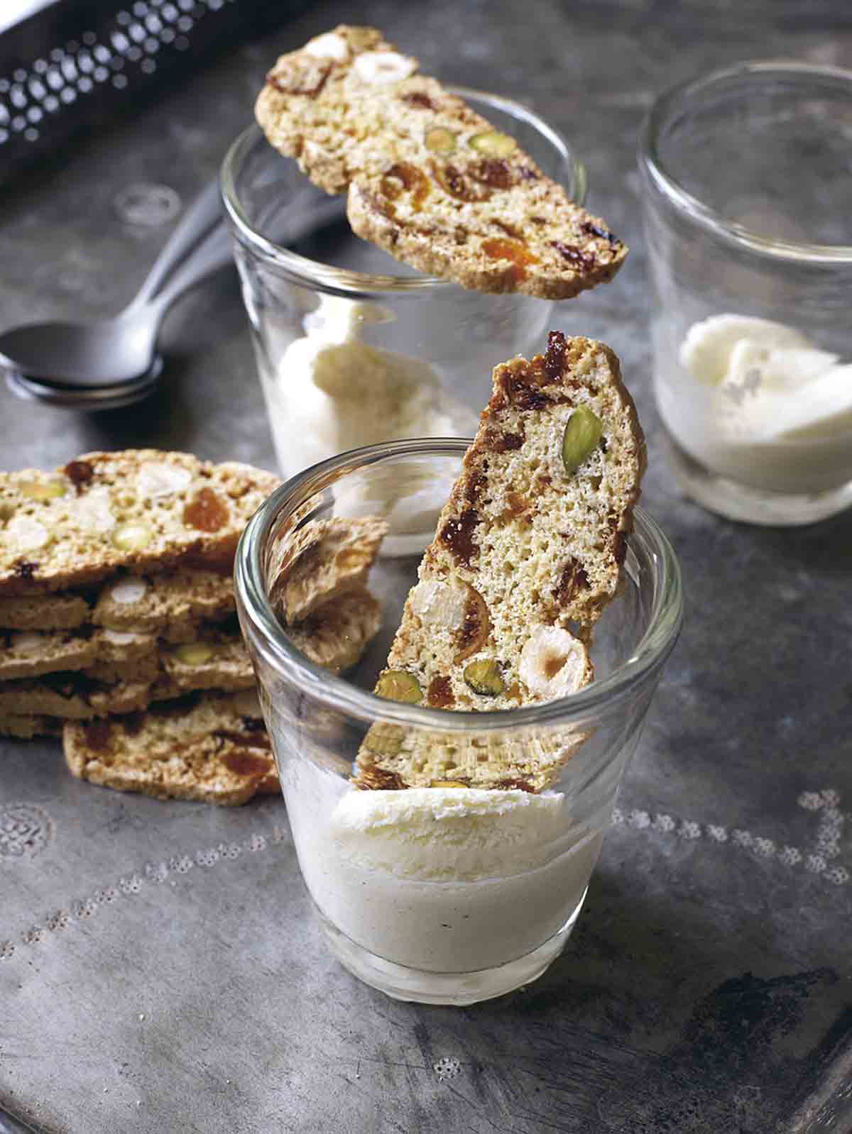 Three glasses with fig, apricot, and pistachio biscotti resting inside or on top of two of them and a stack of biscotti beside the glassware.