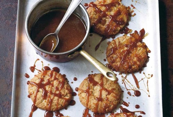 Six ginger-chile caramel cookies on a rimmed baking sheet drizzled with caramel sauce and a pot of caramel sauce.