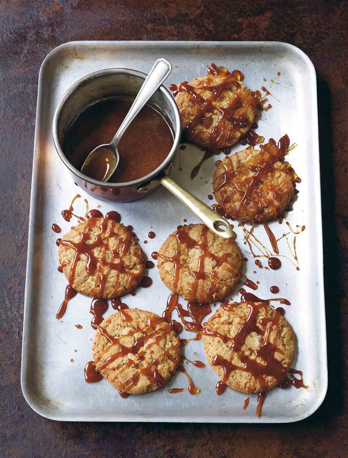 Six ginger-chile caramel cookies on a rimmed baking sheet drizzled with caramel sauce and a pot of caramel sauce.