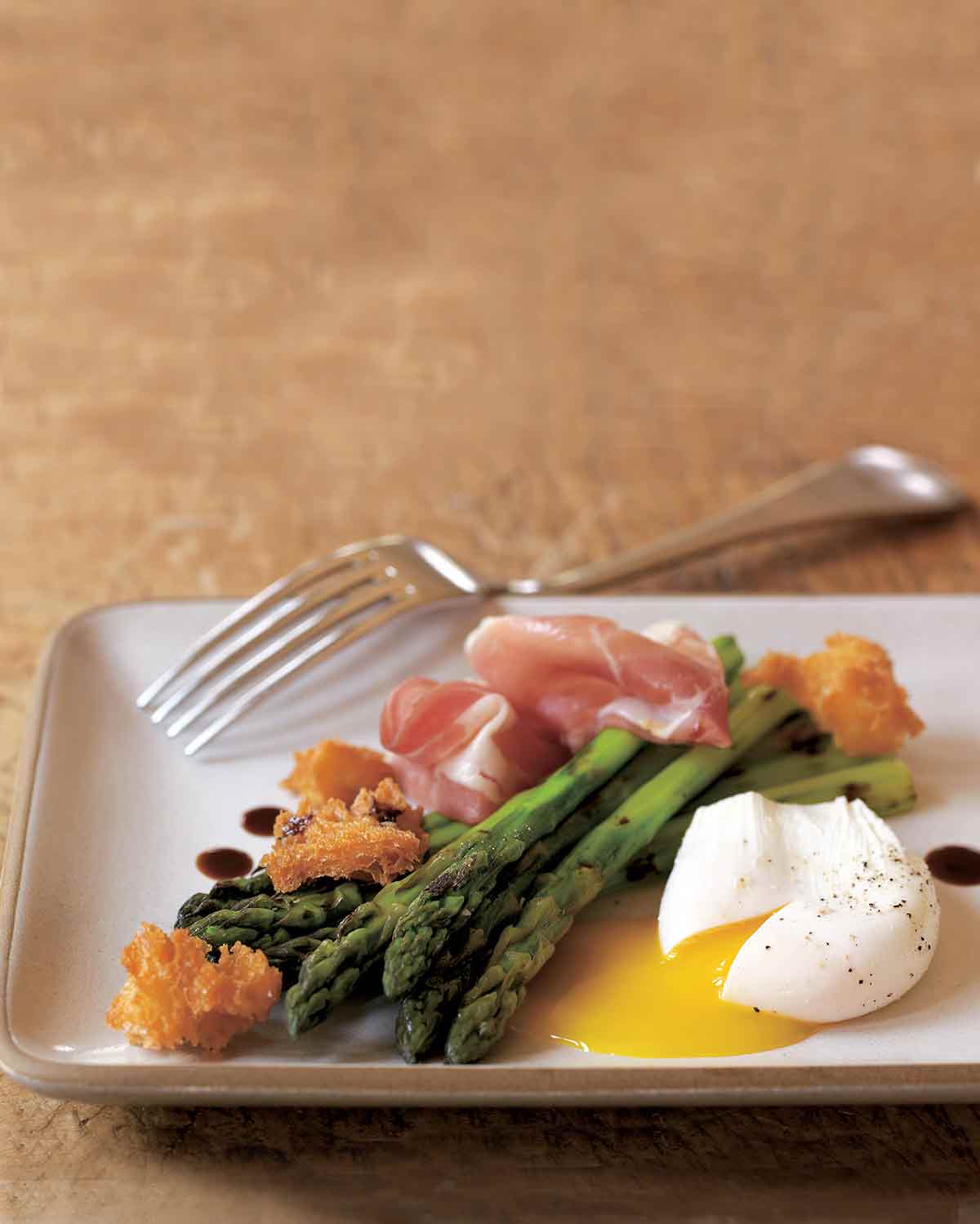 Grilled asparagus, prosciutto, fried bread, and poached egg on a white plate with a fork resting on the side.