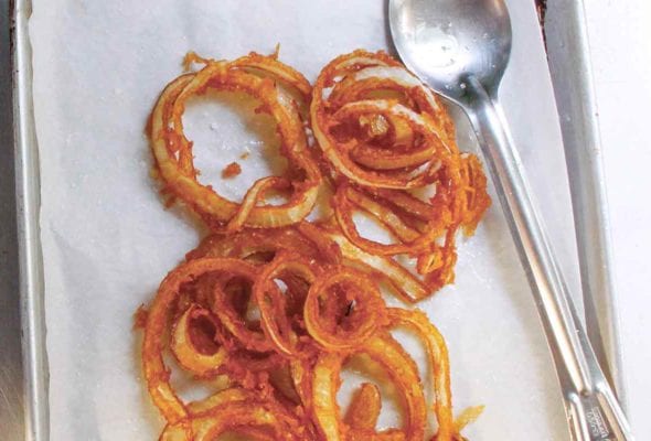 A pile of homemade onion rings on a parchment-lined baking sheet with a spoon resting beside them.