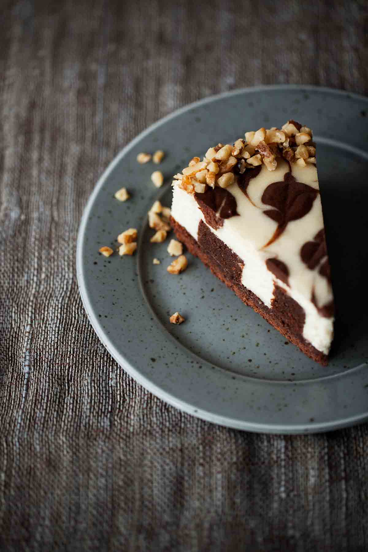 A whole Junior's brownie swirl cheesecake with three slices cut from it, and one of those slices on a grey speckled plate with a fork resting beside.
