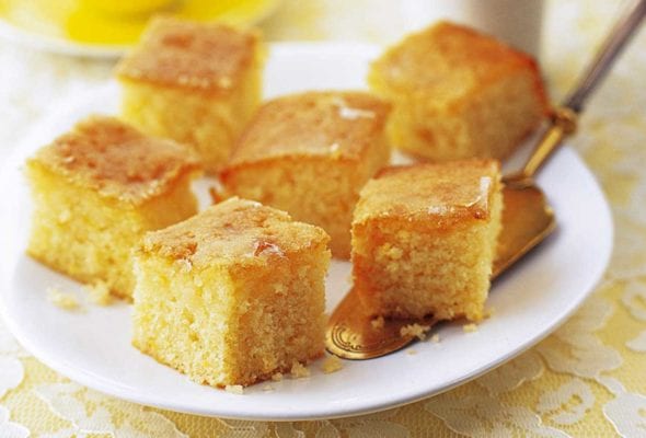 Squares of lemon drizzle cake on a white plate with a cake server resting underneath and a cup of tea and vase of flowers in the background.