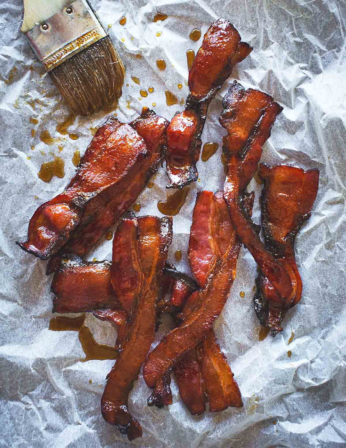 Several strips of maple bacon on a sheet of parchment, with a pastry brush and drizzles of maple syrup beside the bacon.