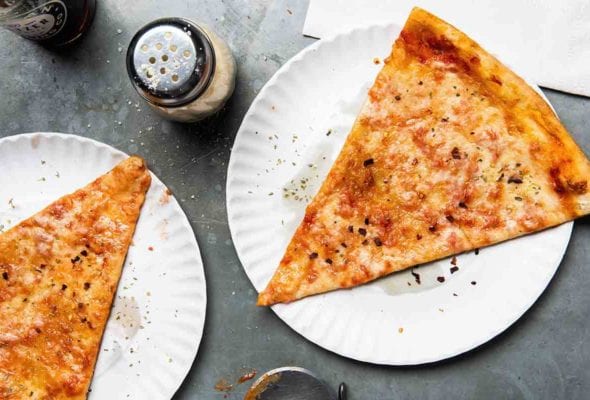 Two slices of New York style pizza on paper plates with a cheese shaker and napkins beside the pizza.