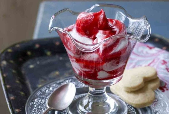 A parfait glass filled with raspberry fool on a decorative glass plate with a silver spoon and two heart-shaped cookies.