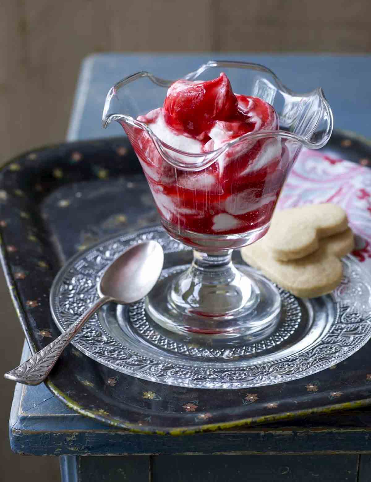 A parfait glass filled with raspberry fool on a decorative glass plate with a silver spoon and two heart-shaped cookies.