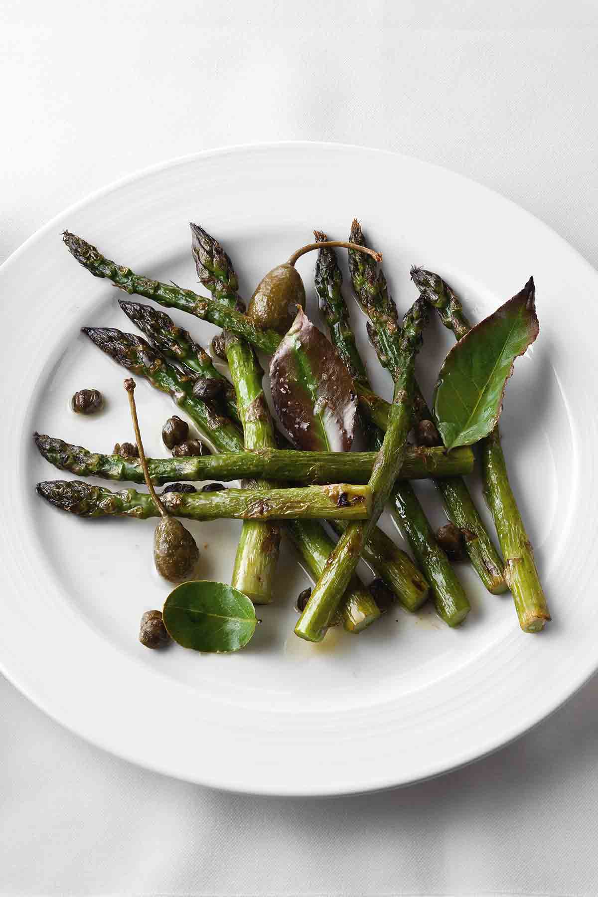 A white plate topped with roasted asparagus with bay leaves and crispy capers.