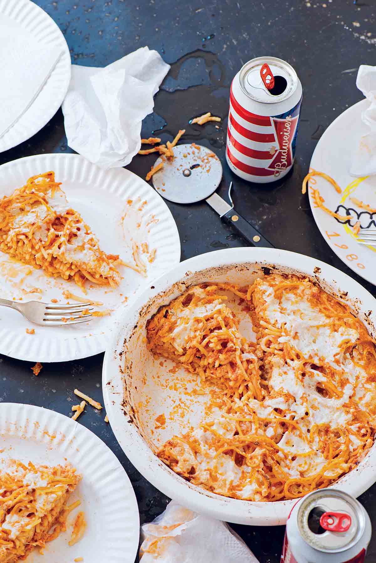 A white dish with a partially cut spaghetti pie, two paper plates with slices of spaghetti pie, a pizza cutter, napkins, and two cans of beer.