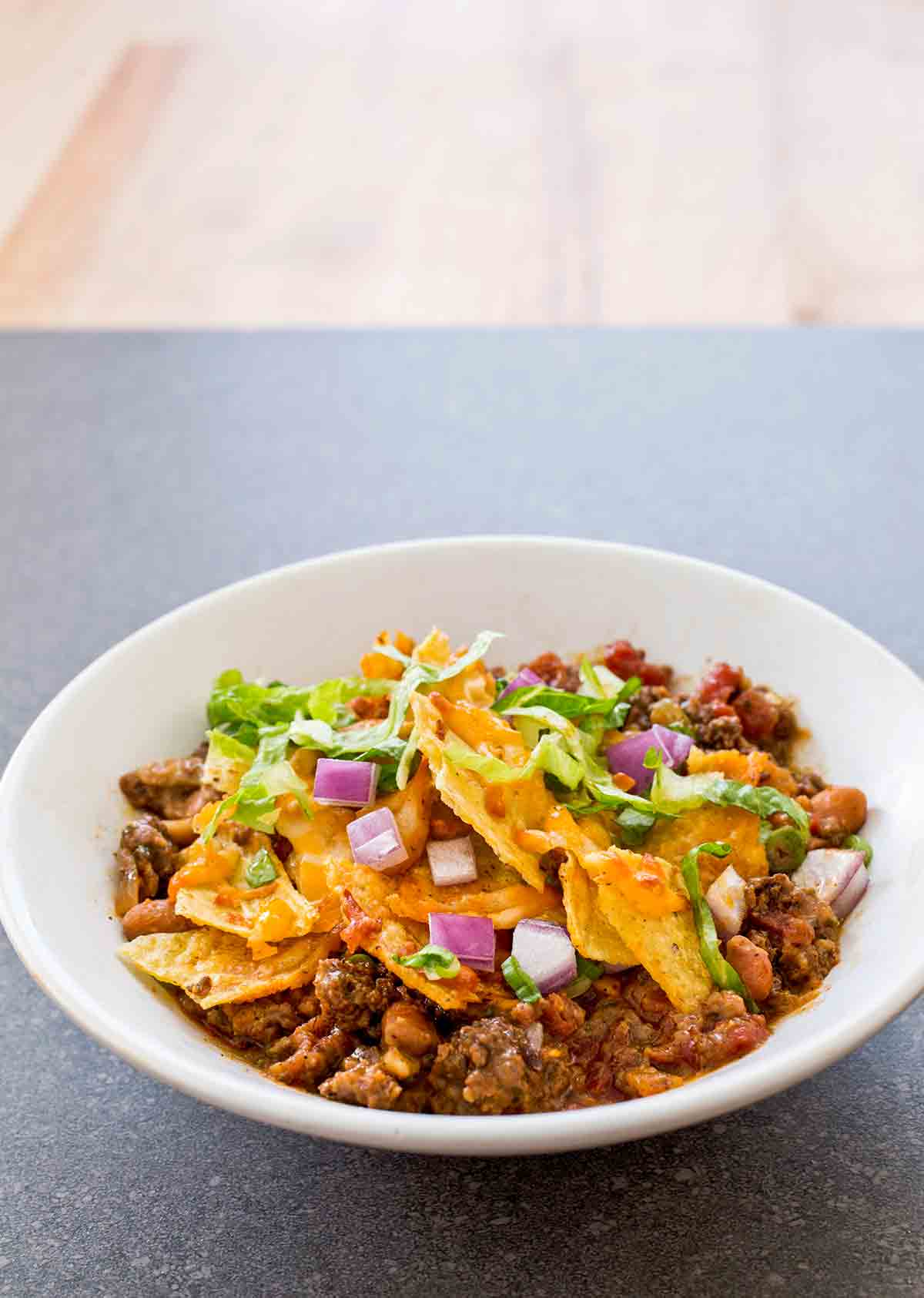 White bowl of spicy beef taco bake with ground beef, beans tortilla chips, chopped onions, and lettuce