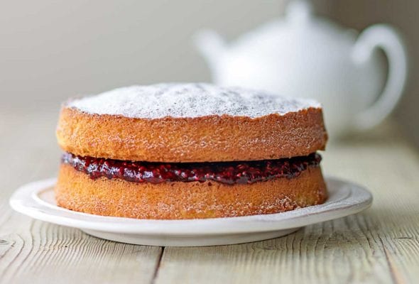 A two-tier Victoria sponge cake with raspberry jam in the middle, all dusted with confectioners' sugar on a white plate with a white teapot in the background