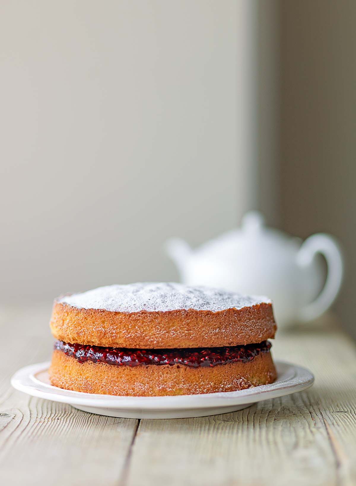 A two-tier Victoria sponge cake with raspberry jam in the middle, all dusted with confectioners' sugar on a white plate with a white teapot in the background