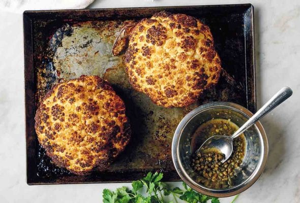 Two whole roasted cauliflower heads on a rimmed baking sheet with a glass jar of dressing and a spoon resting inside it.