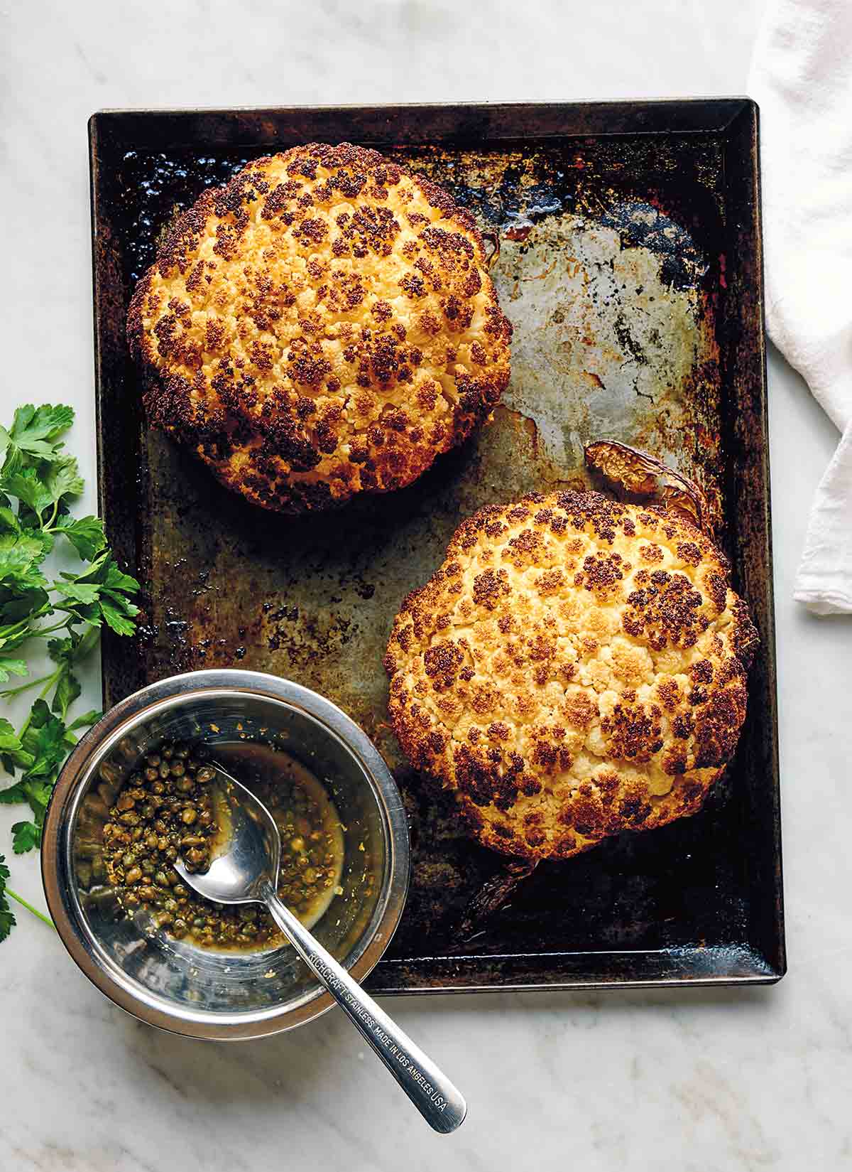 Two whole roasted cauliflower heads on a rimmed baking sheet with a glass jar of dressing and a spoon resting inside it.