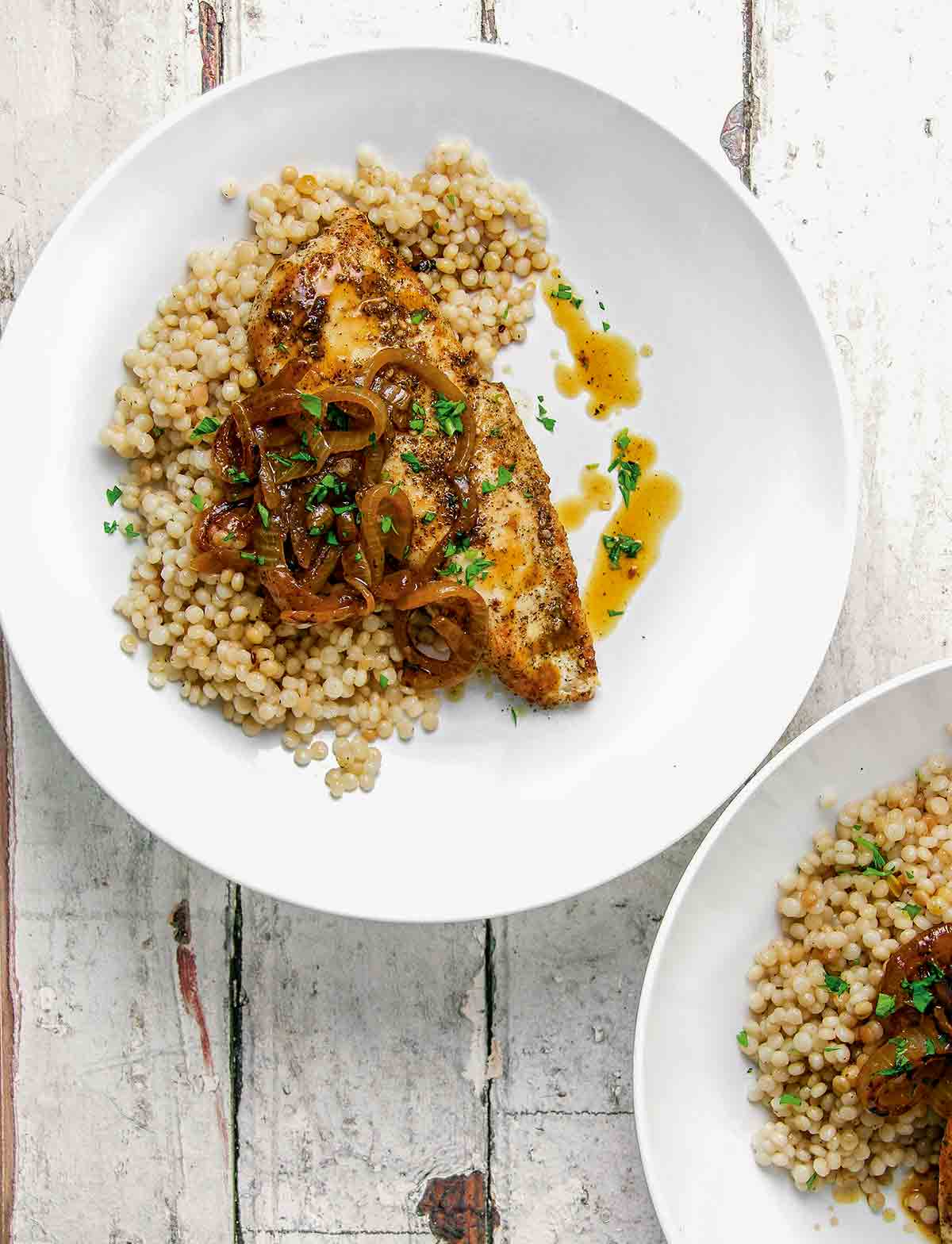 Two white plates each topped with a serving of za'atar chicken, Israeli couscous, sauteed onions, and pan juices.