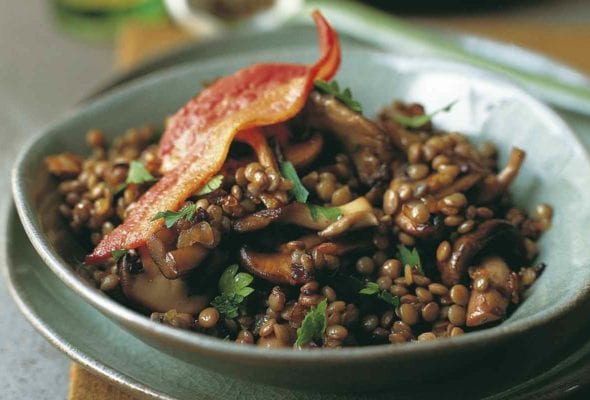 A green bowl filled with sautéed bacon, mushrooms, and lentils with a whole piece of bacon on top.