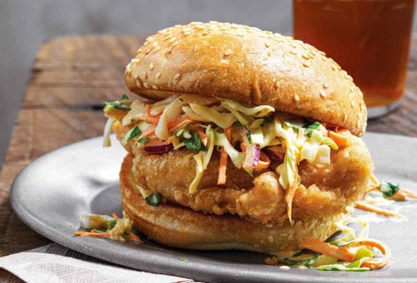 A beer battered chicken sandwich topped with slaw on a white plate on top of a paper napkin on a wooden table with a glass of beer in the background.