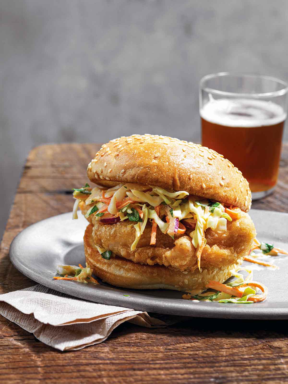 A beer battered chicken sandwich topped with slaw on a white plate on top of a paper napkin on a wooden table with a glass of beer in the background.