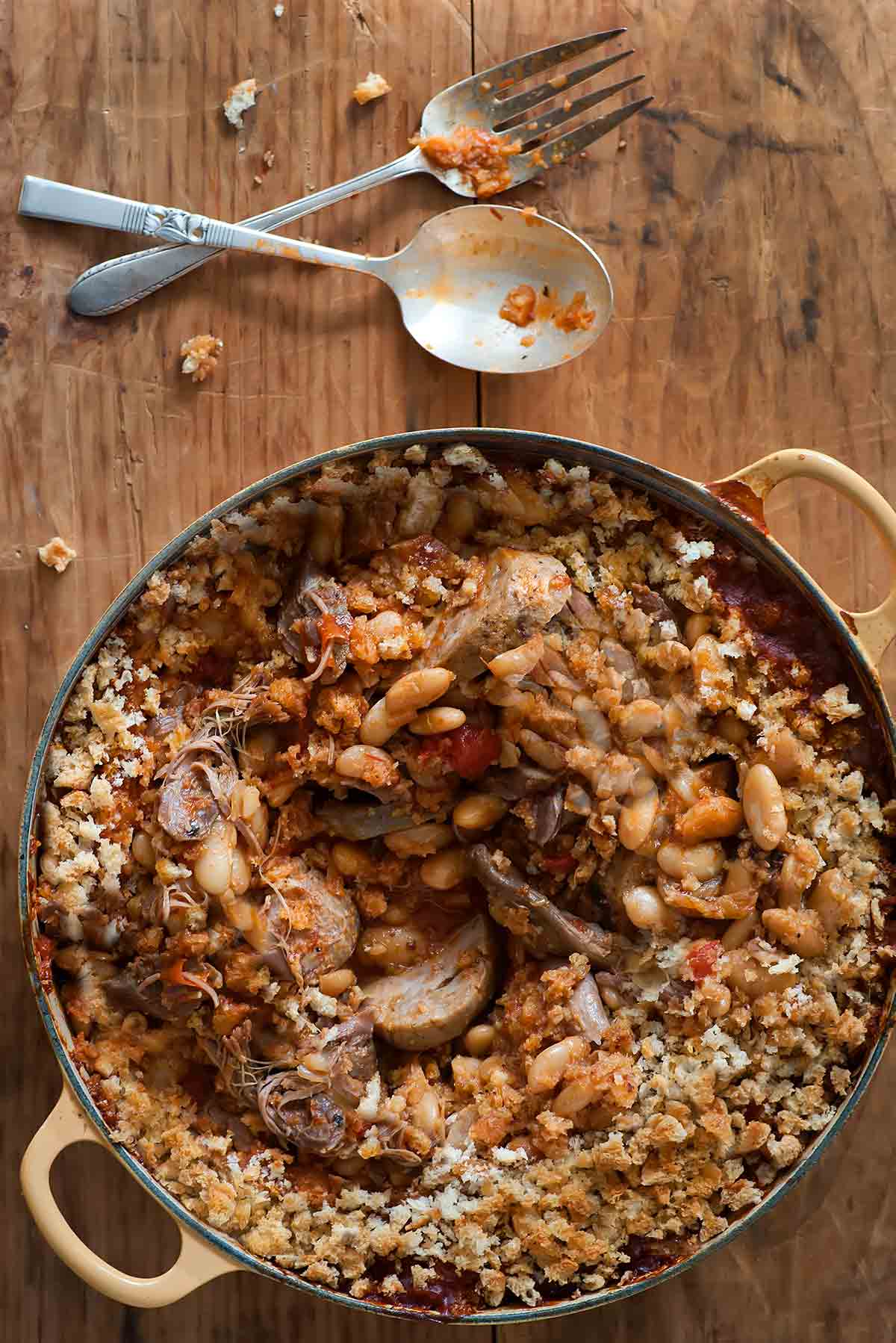 A cassoulet of white beans, sausage, and duck in a yellow Dutch oven with a spoon and fork resting beside it on a wooden surface.