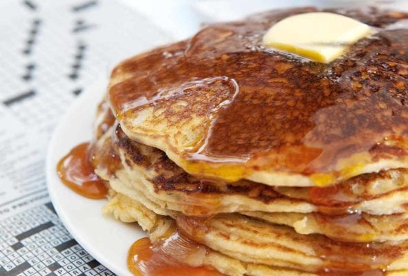 A stack of gluten-free pancakes with butter and maple syrup on a white plate on top of a crossword puzzle.