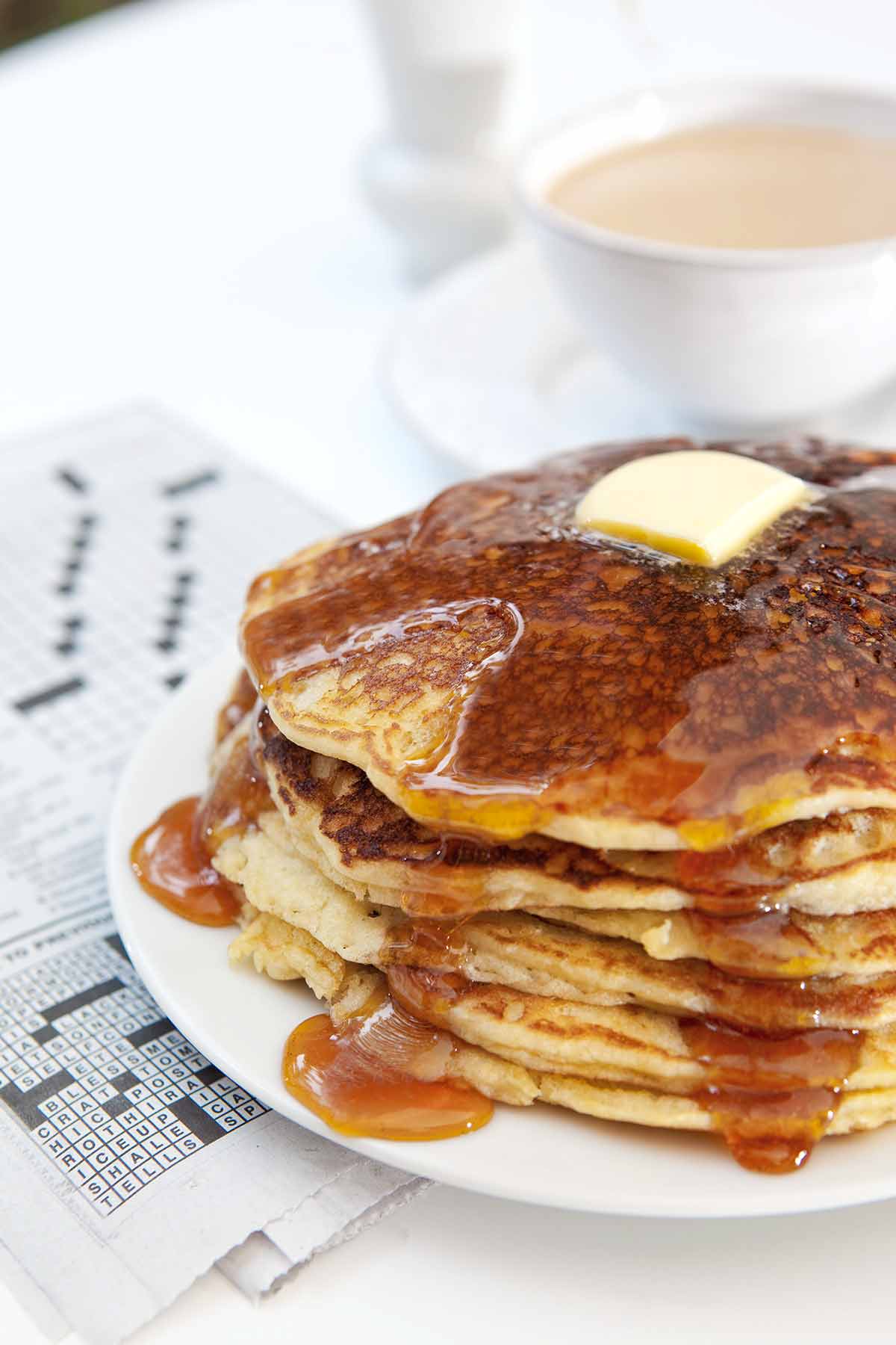 A stack of gluten-free pancakes with butter and maple syrup on a white plate on top of a crossword puzzle.