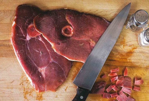 A slice of ham with red eye gravy on a cutting board with a knife and salt and pepper shaker.