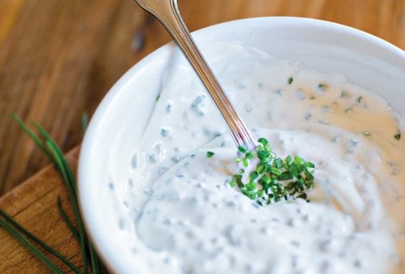 A bowl of horseradish cream on a wooden board with a spoon resting inside and a bunch of chives lying beside the bowl.