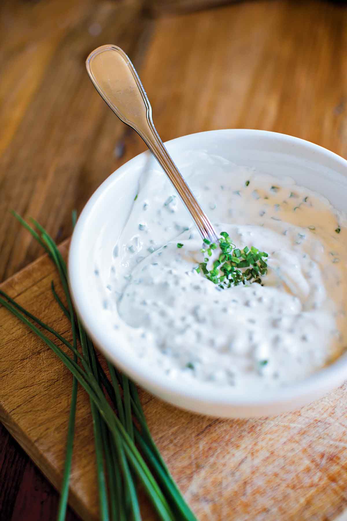 A bowl of horseradish cream on a wooden board with a spoon resting inside and a bunch of chives lying beside the bowl.