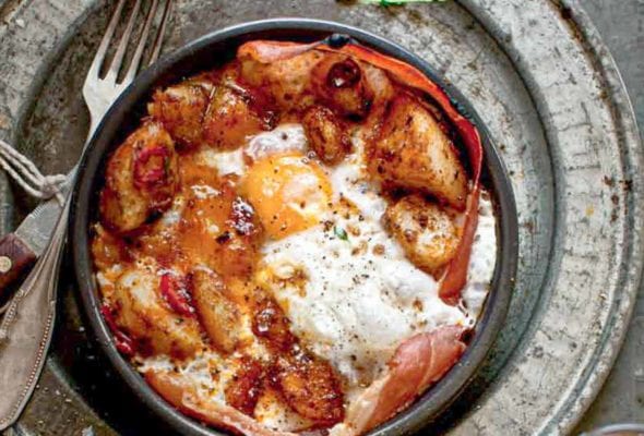 A bowl of patatas bravas with ham and egg - crispy potatoes, ham, and fried egg - with a fork resting beside it.