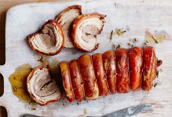 A pork belly porchetta on a cutting board with four slices carved from the end and a carving fork and knife resting beside the meat.