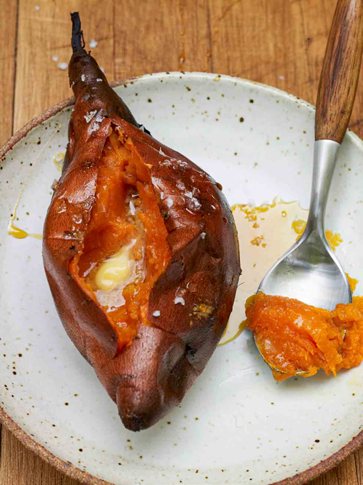 A roasted sweet potato on a speckled plate with a dollop of butter melting into the flesh and a small spoon with a scoop of potato on it resting beside the whole sweet potato.