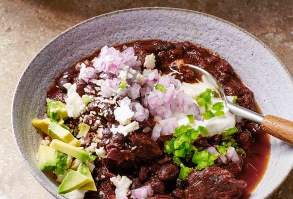 A white bowl filled with beef and black bean chili, topped with red onion, avocado, queso fresco, and diced jalapeno
