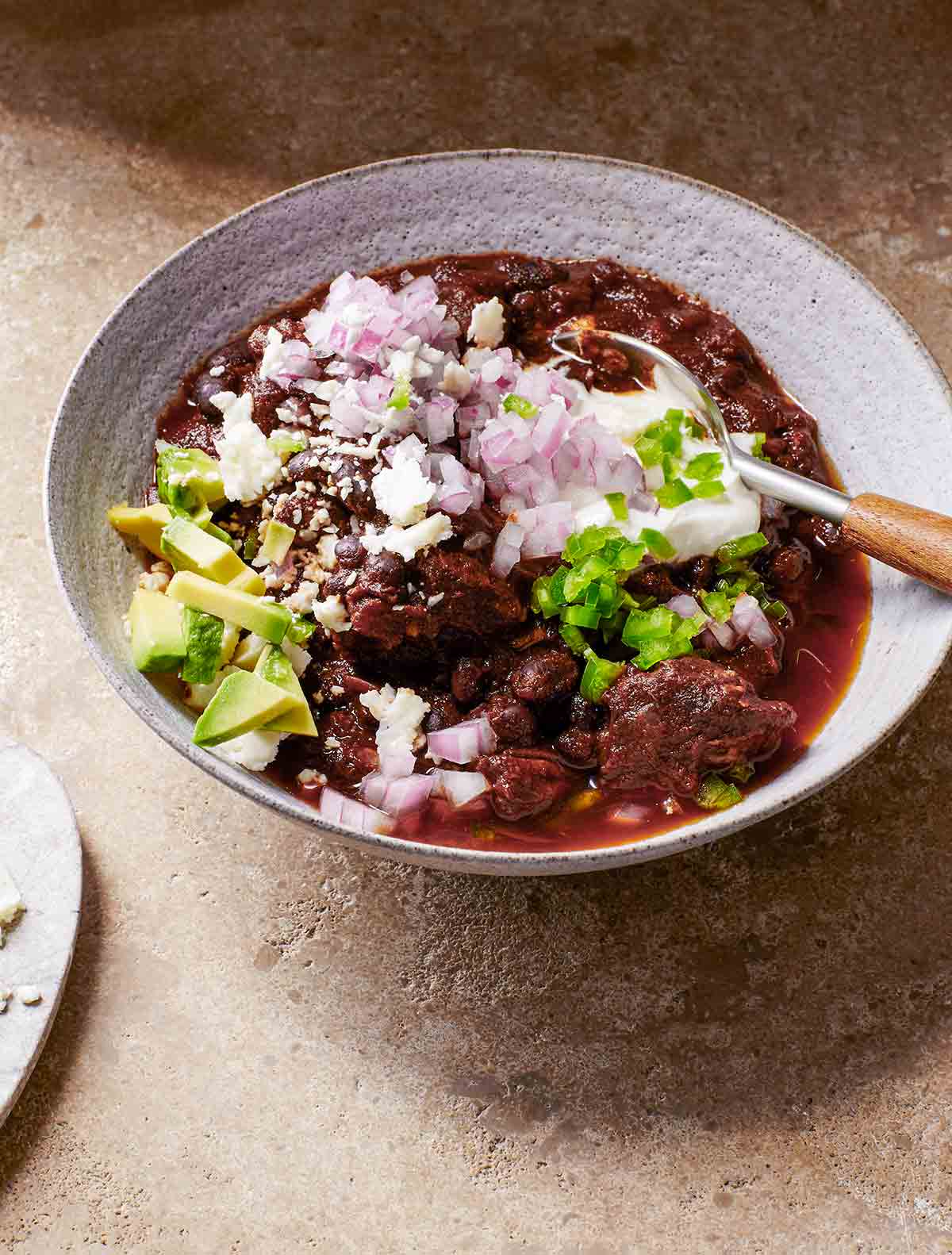 A white bowl filled with beef and black bean chili, topped with red onion, avocado, queso fresco, and diced jalapeno