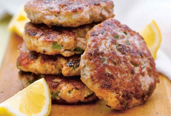 Five turkey burgers with lemon and mint stacked on a wooden board with lemon wedges.